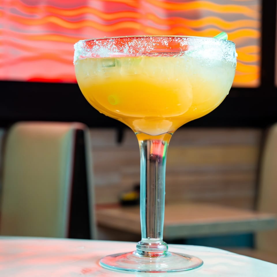 Close-up of a vibrant yellow margarita with a salted rim and green straw, on a table at Mama's Tacos Delicioso.