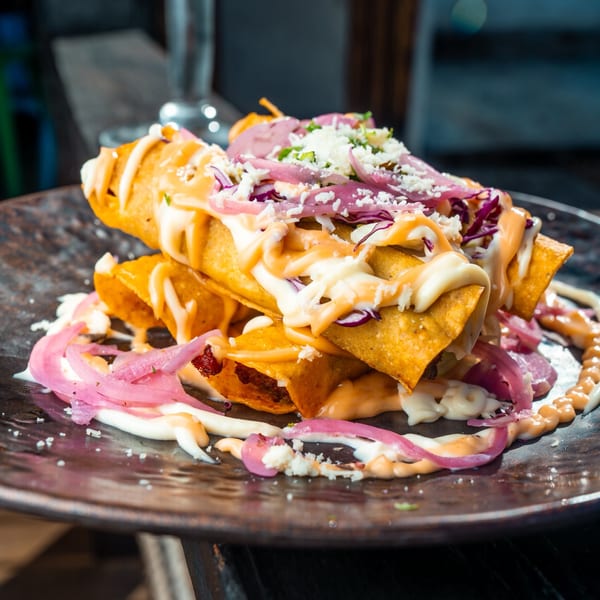 Crispy taquitos from Mama's Tacos Delicioso, topped with pickled red onions, cheese, and sauces.
