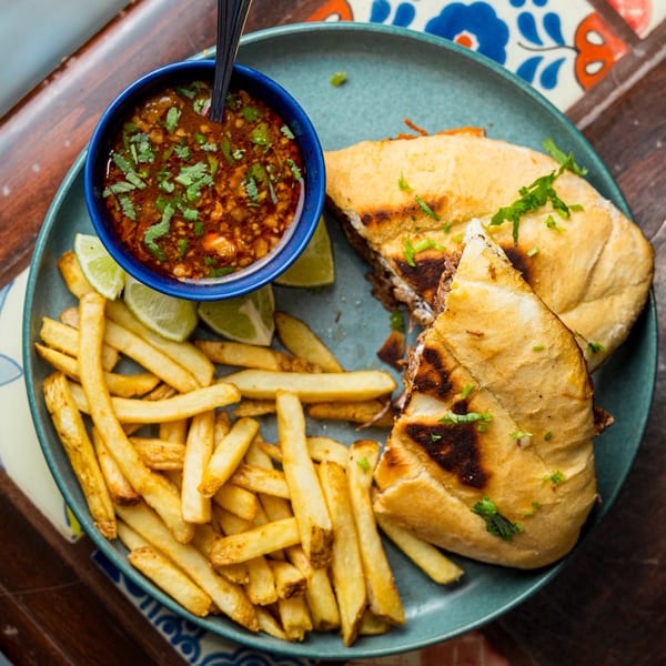 A Mexican torta sandwich, fries, and a bowl of birria broth with lime wedges on a blue plate.