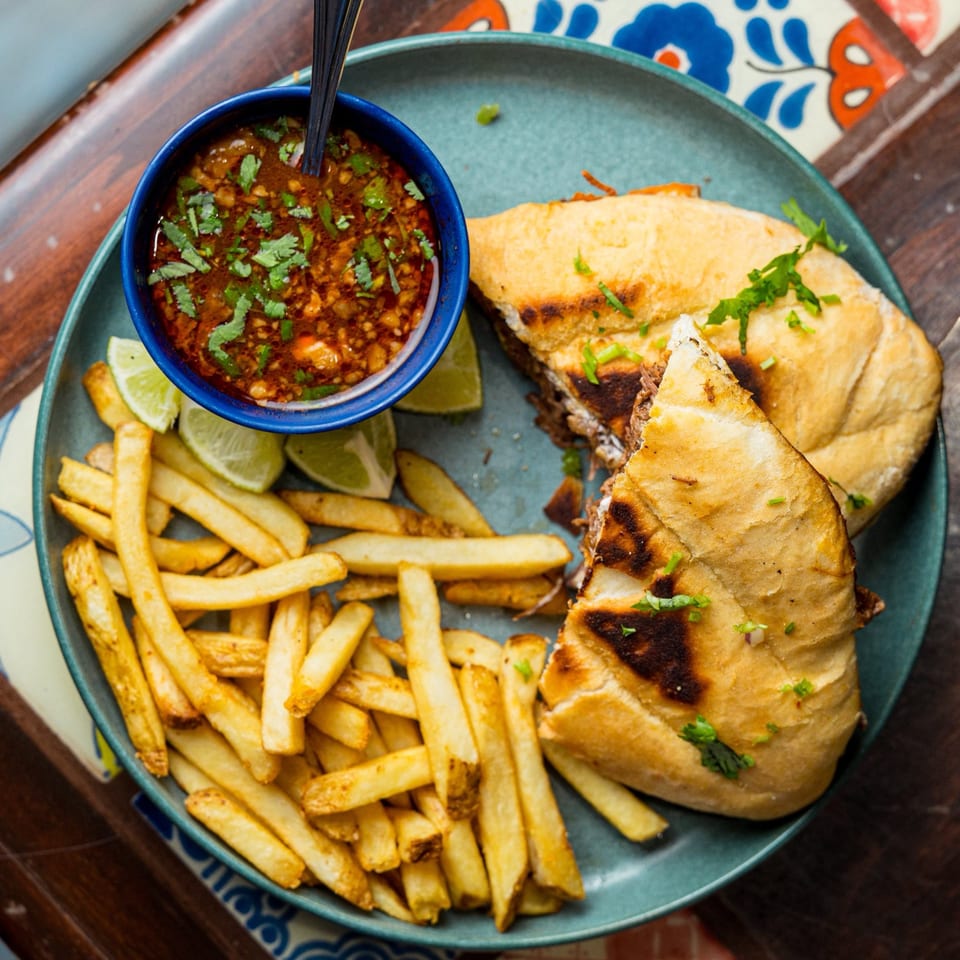 A Mexican torta sandwich, fries, and a bowl of birria broth with lime wedges on a blue plate.