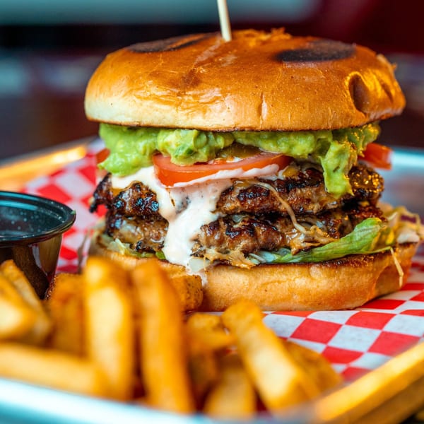A large double-patty burger with guacamole, tomatoes, and white sauce, served with fries.