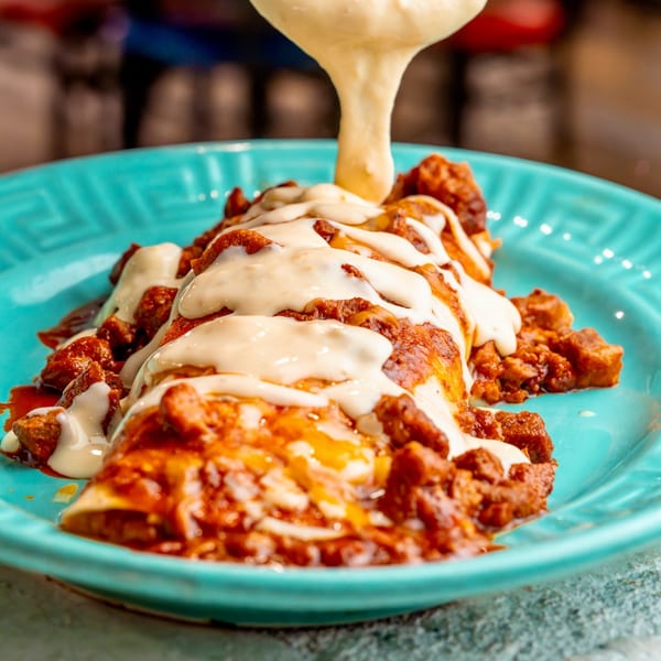 Creamy white queso sauce being poured over a large, saucy burrito on a turquoise plate.