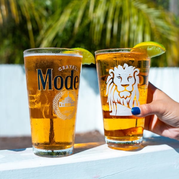 Two Modelo beers with lime wedges, one held by a hand with blue nail polish, outdoors.