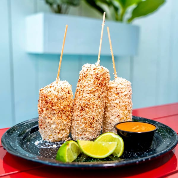Three elotes, Mexican street corn, on sticks with lime wedges and a side of sauce.