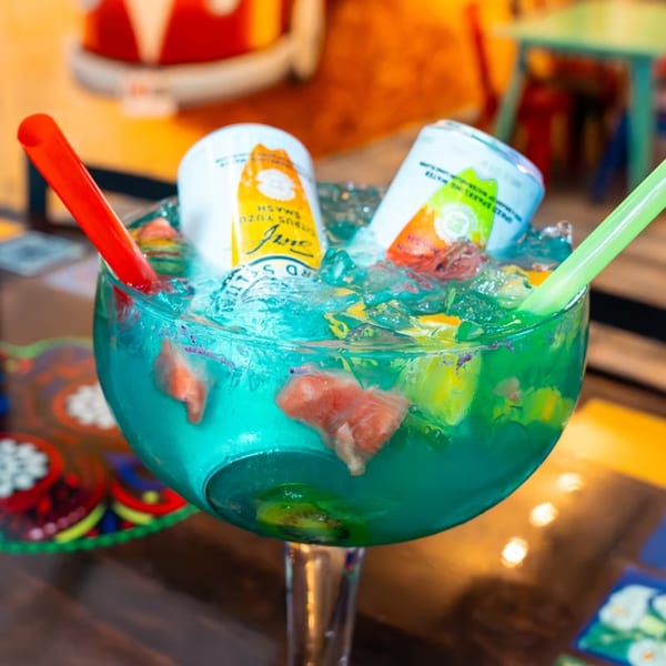 Giant blue margarita with two cans, ice, fruit, and colorful straws at Mama's Tacos Delicioso.